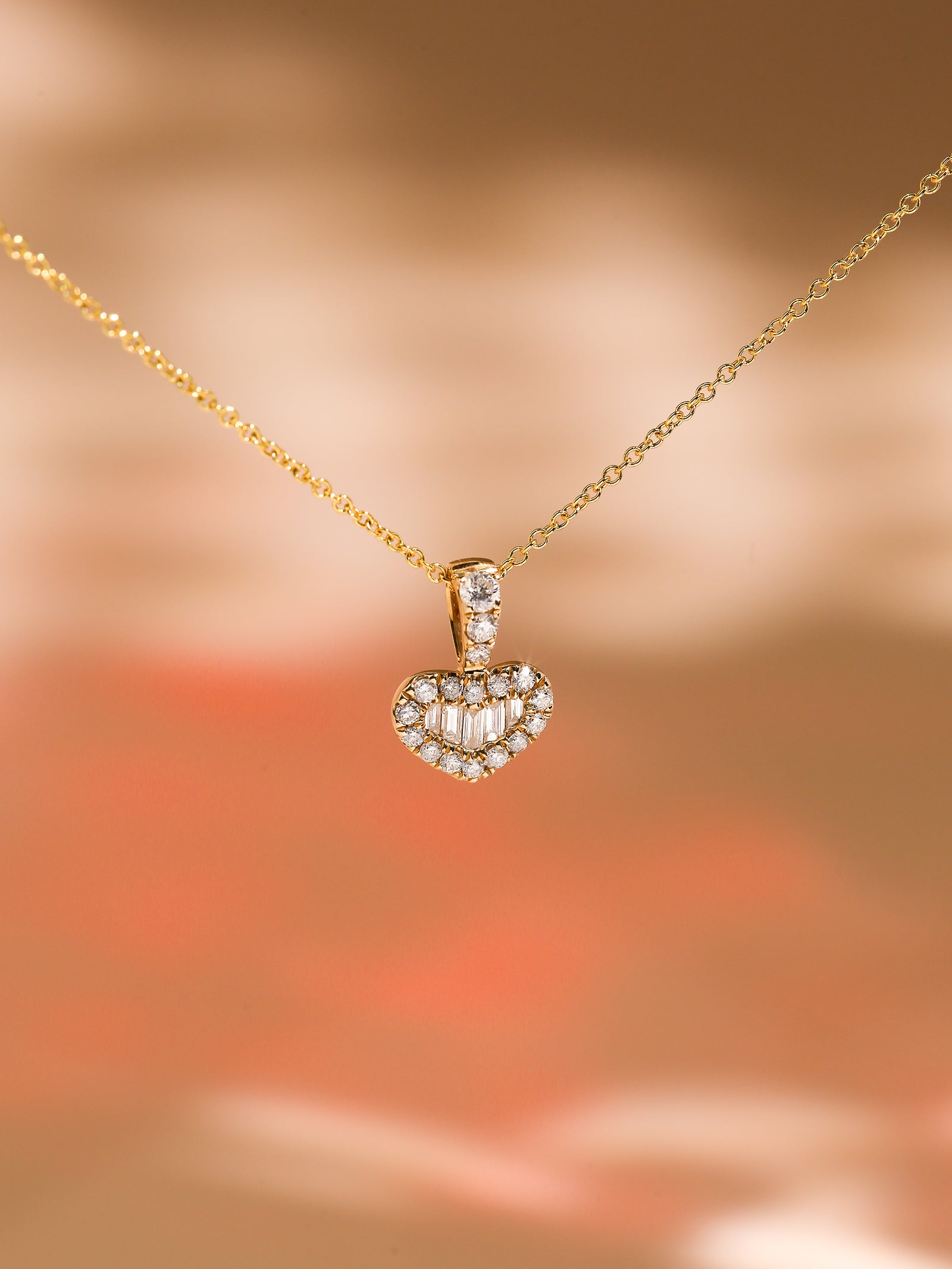 A Kobelli heart-shaped diamond pendant on a gold chain against a blurred warm background