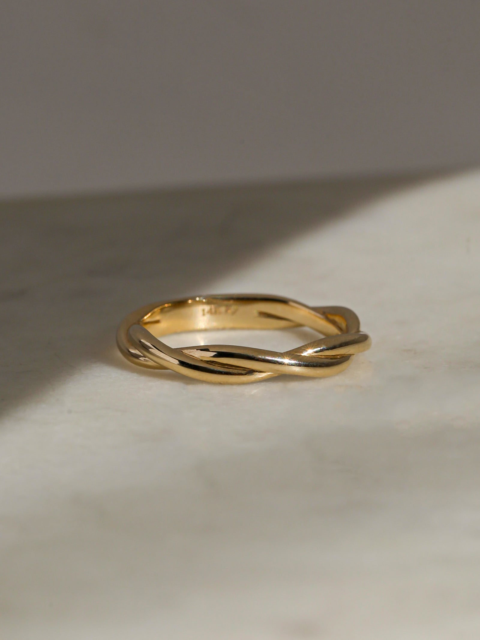 Twisted yellow gold band resting on a light marble surface with soft shadows.