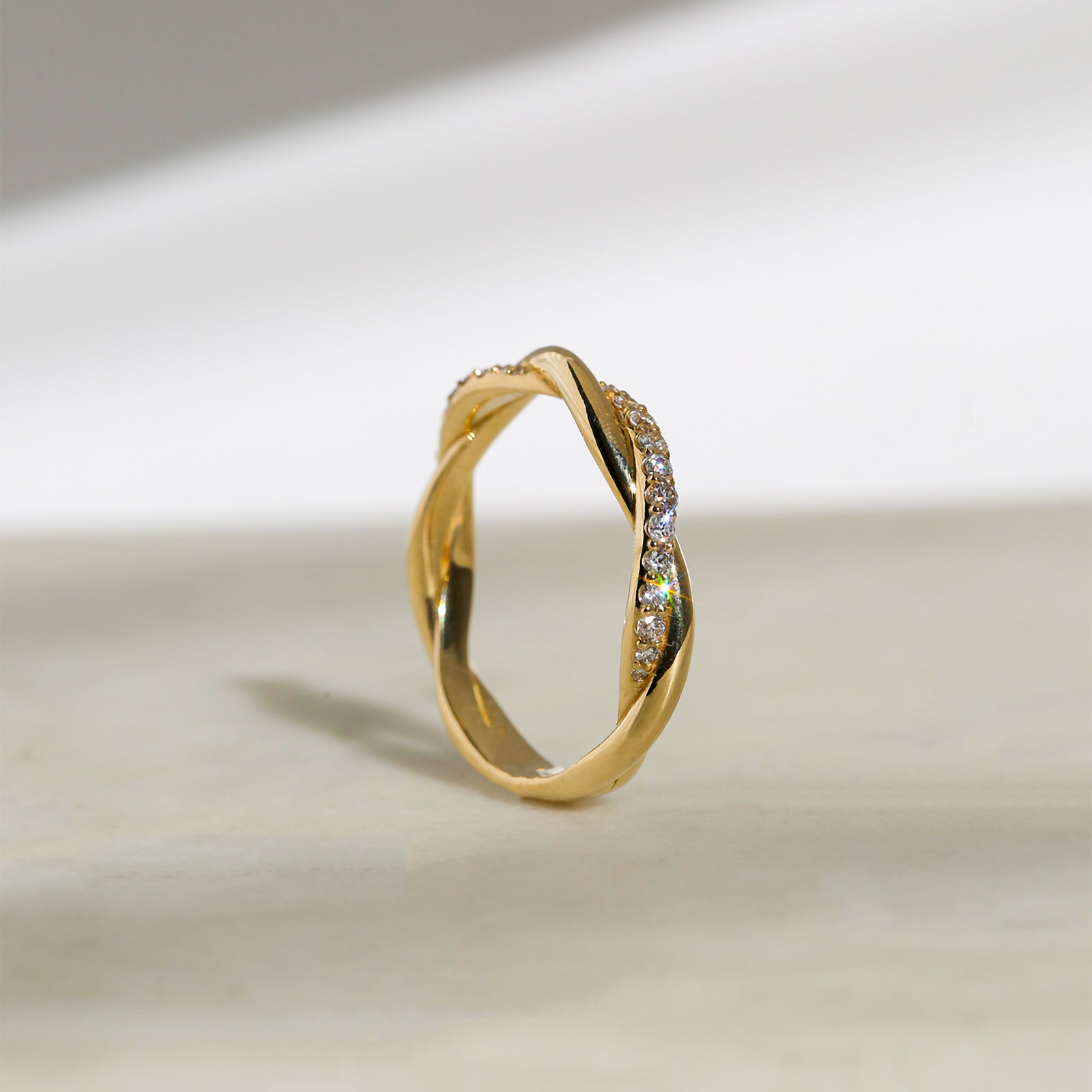 Yellow gold twist band ring with a row of diamonds on one side, vertical tilted angle to showcase the design of the entire band. Photographed on a light beige surface with a soft shadow.