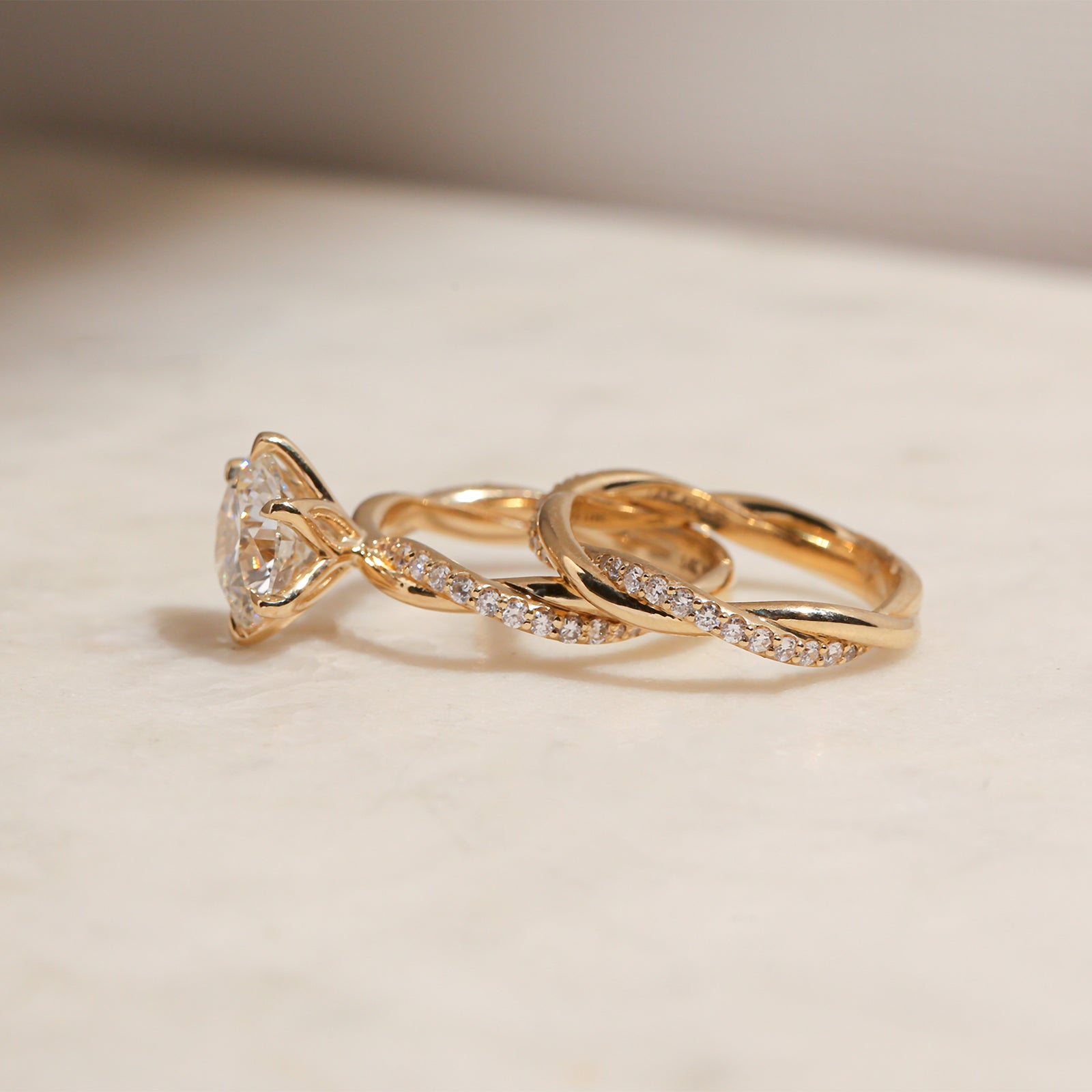side angle of the 3.25 Carat Lab Grown Diamond Round Shifting Helix Ring paired with the Shifting Helix Wedding Band Ring, leaning against the bottom edge of the engagement ring - yellow gold option resting on an off-white table with subdued daylight
