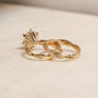 back angle of the 3.25 Carat Lab Grown Diamond Round Shifting Helix Ring paired with the Shifting Helix Wedding Band Ring, leaning against the bottom edge of the engagement ring - yellow gold option resting on an off-white table with subdued daylight
