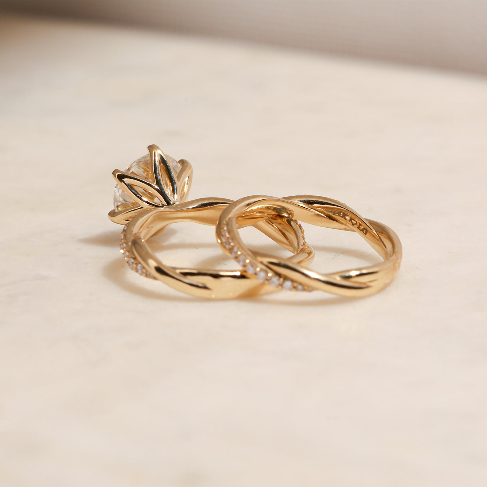 back angle of the 3.25 Carat Lab Grown Diamond Round Shifting Helix Ring paired with the Shifting Helix Wedding Band Ring, leaning against the bottom edge of the engagement ring - yellow gold option resting on an off-white table with subdued daylight