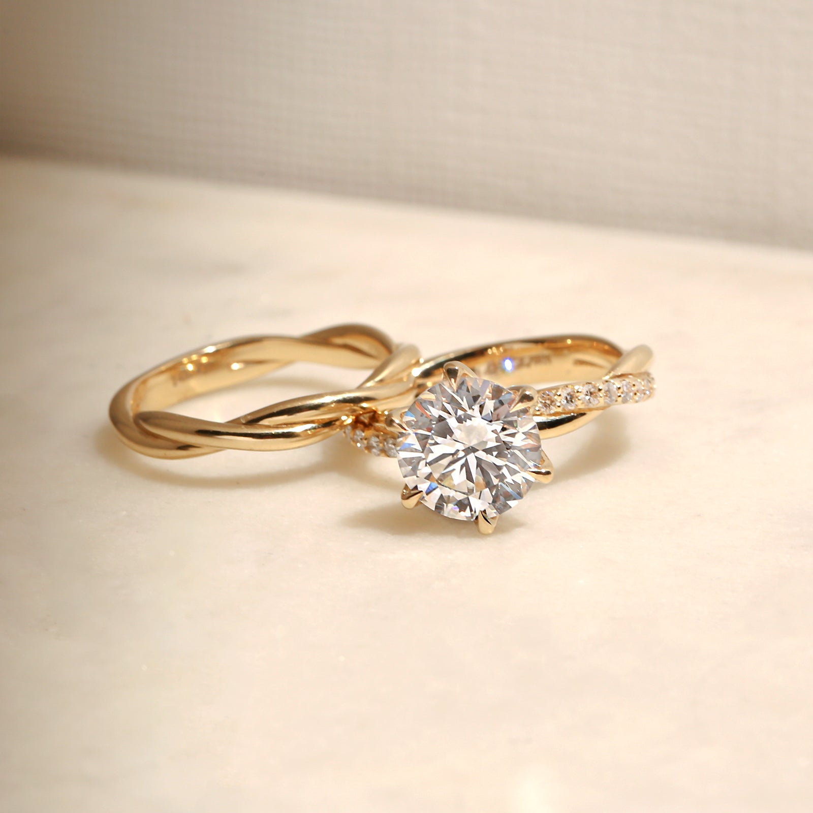 front angle of the 3.25 Carat Lab Grown Diamond Round Shifting Helix Ring paired with the Plain Helix Wedding Band Ring, leaning against the bottom edge of the engagement ring - yellow gold option resting on an off-white table with subdued daylight
