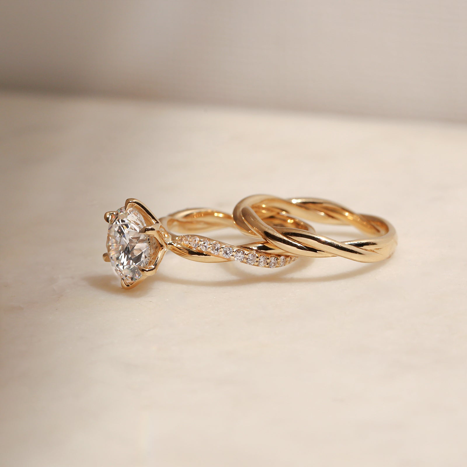 side angle of the 3.25 Carat Lab Grown Diamond Round Shifting Helix Ring paired with the Plain Helix Wedding Band Ring, leaning against the bottom edge of the engagement ring - yellow gold option resting on an off-white table with subdued daylight