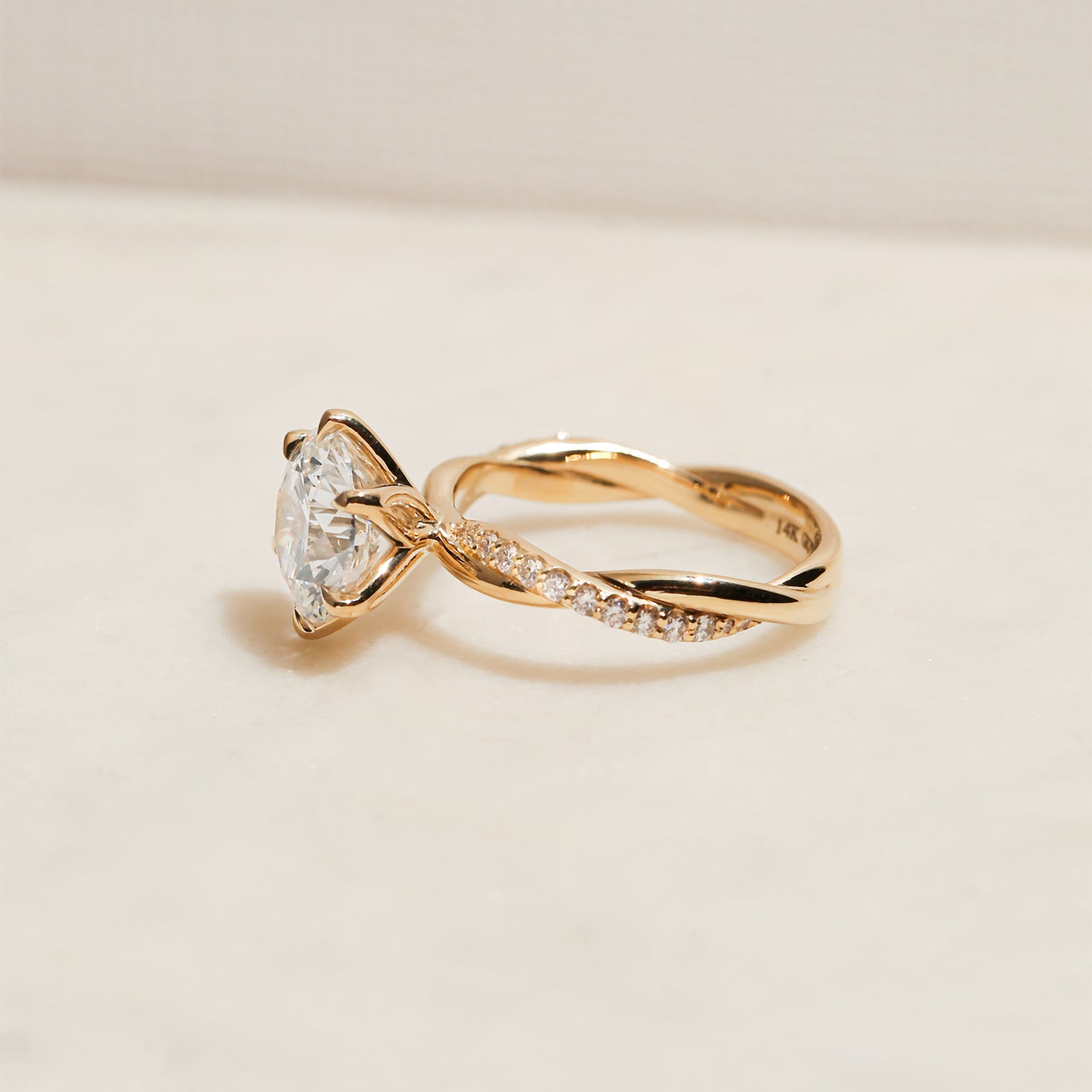 3.25 Carat Lab Grown Diamond Round Shifting Helix Ring - lifestyle image of yellow gold option resting on an off-white table with subdued daylight, photographed at a side angle slightly above the ring to show depth and details