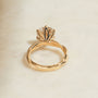 Yellow gold ring with a lotus flower-inspired setting and a clear central gemstone, resting on a light beige surface.