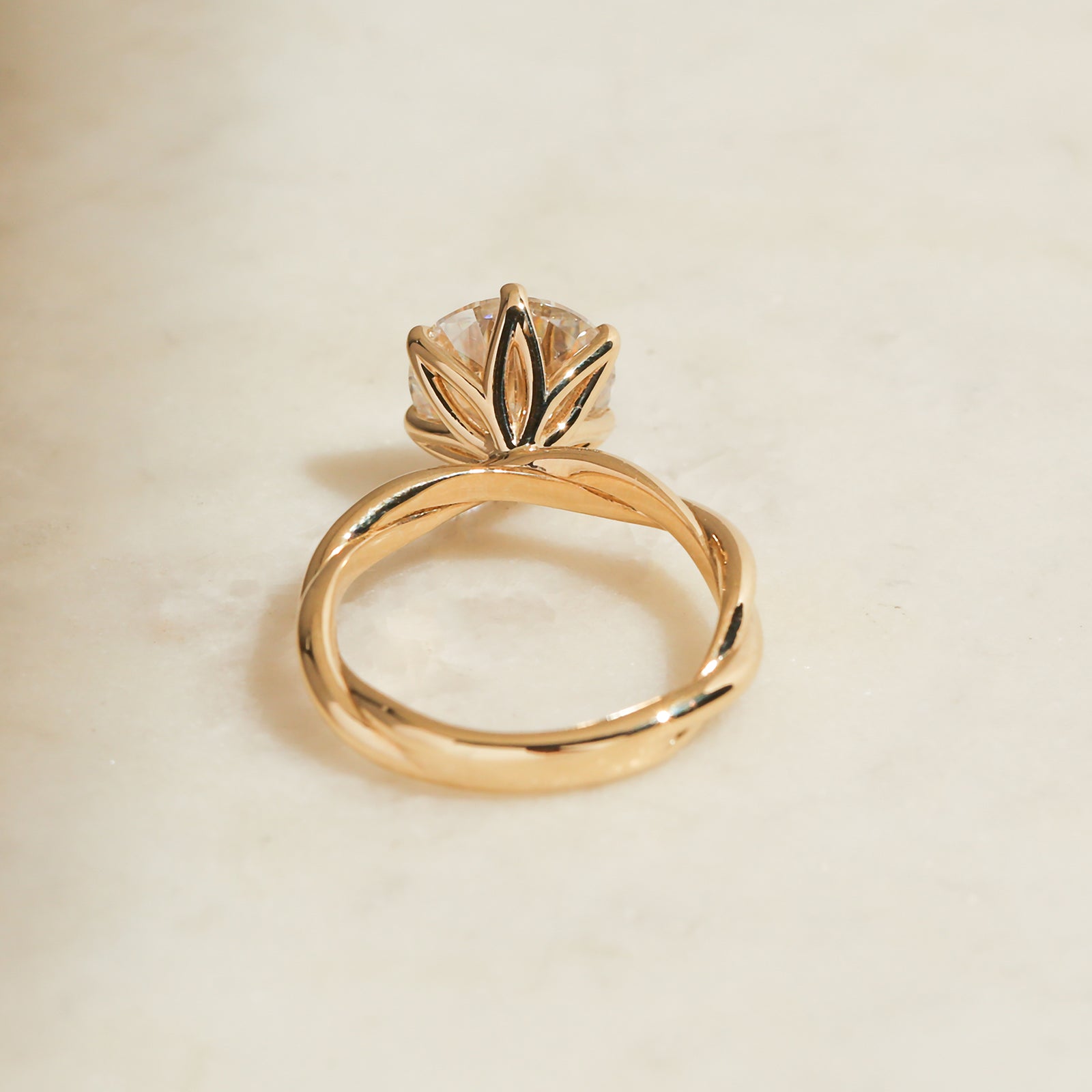 Yellow gold ring with a lotus flower-inspired setting and a clear central gemstone, resting on a light beige surface.