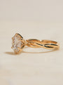 Yellow gold ring with a twisted band and a large clear round gemstone in a prong setting, photographed on a plain light beige background.