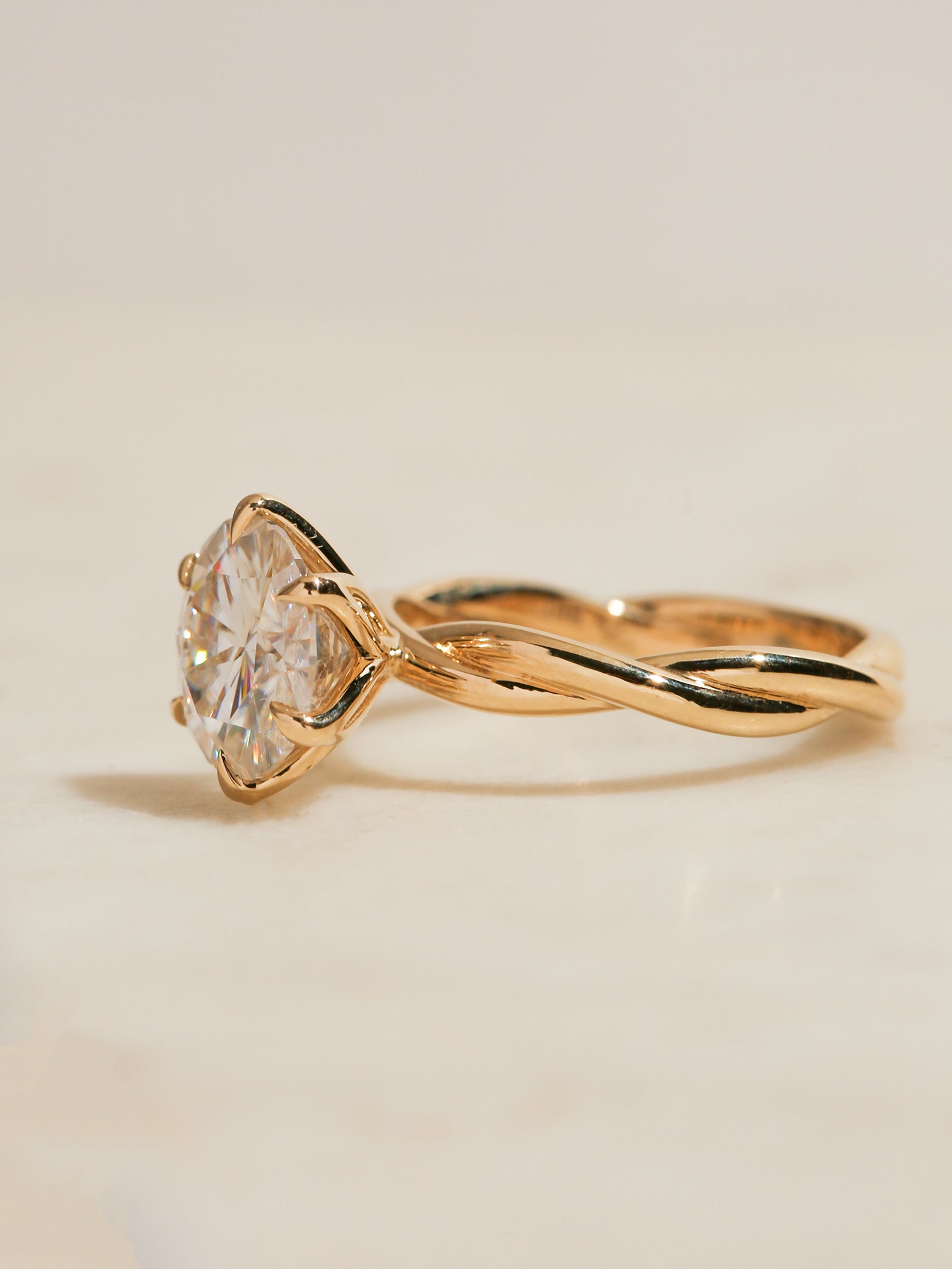 Yellow gold ring with a twisted band and a large clear round gemstone in a prong setting, photographed on a plain light beige background.