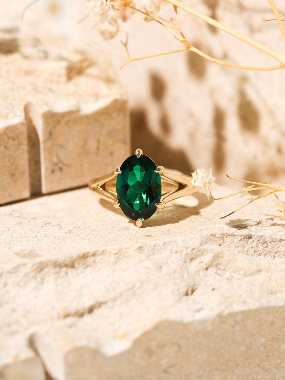 A custom Kobelli Ring featuring a long oval green gemstone set with 6 prongs in a split shank solid yellow gold band - lifestyle shot with the ring sitting in warm sandy stone scenery and dried desert branches